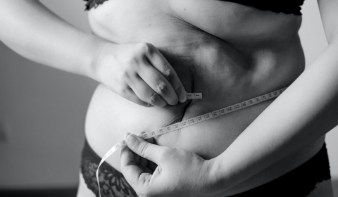 A black and white image of a person measuring their waistline with a tape measure.