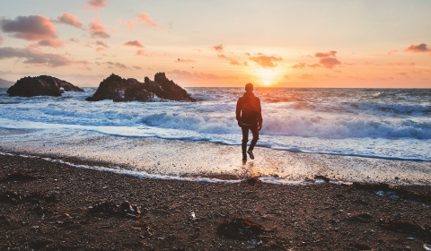 A person stood ankle-deep in the sea, looking out at sunset over the waves. An image by Joshua Earle on Unsplash.
