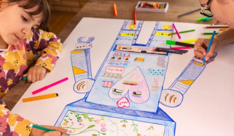 Two children drawing a robot on paper with crayons