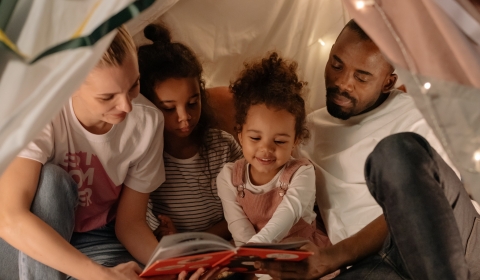 A happy family of two adults and two children reading a bedtime story in a den decorated with fairy lights
