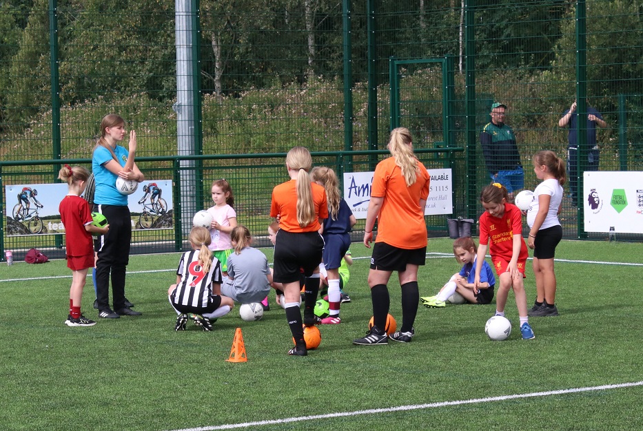 Female football coach coaching around a dozen girls in football strips in a group with footballs